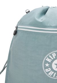 Sacca azzurra chiara con cordino, dotata di una tasca con zip bianca e un logo rotondo parziale bianco con stella e testo sul tessuto.