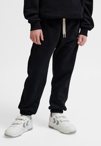 Schwarze Jogginghosen mit elastischen Bündchen und einem Kordelzugbund, kombiniert mit weißen Sneakers mit grauen Akzenten und Klettverschlüssen.