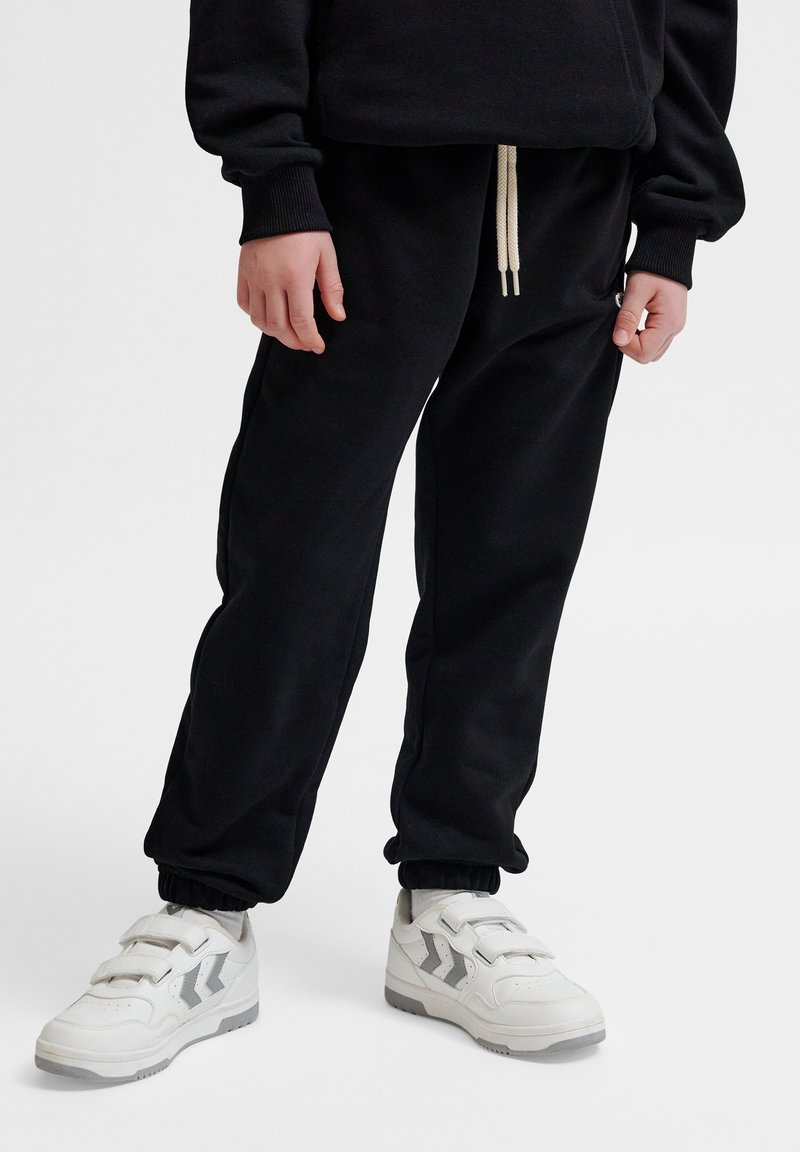 Schwarze Jogginghosen mit elastischen Bündchen und einem Kordelzugbund, kombiniert mit weißen Sneakers mit grauen Akzenten und Klettverschlüssen.