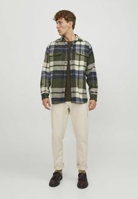 Groene en roomkleurige geblokte flanellen shirt met voorzakken, knoopsluiting; gecombineerd met beige broek en donkere schoenen.