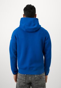 Μπλε φούτερ με μεγάλη κουκούλα και κορδόνι, με ραμμένα μανίκια και κάτω μέρος. Διαθέτει λεία υφή και ελάχιστες ραφές. Φοριέται με γκρι τζιν.