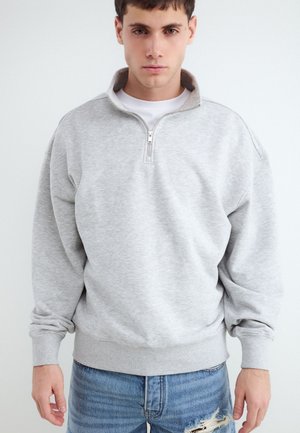 Jonge man met een lichtgrijze sweatshirt met een kwart rits en gescheurde blauwe spijkerbroek, staand tegen een effen witte achtergrond.