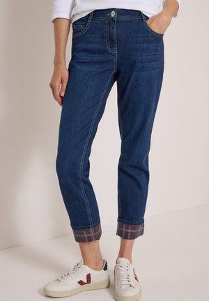 Femme portant un jean bleu avec revers et doublure à carreaux, ainsi que des baskets blanches avec des touches rouges, debout avec une main dans la poche.