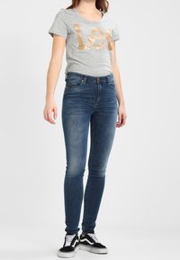 Camiseta de manga corta gris con el texto metálico dorado "Let", combinada con jeans ajustados azul oscuro y zapatillas en blanco y negro.