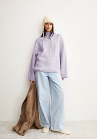 Lila kabelgestrickter Pullover mit hohem Kragen, kombiniert mit hellblauen weit geschnittenen Jeans. Ein brauner Mantel über dem Arm und cremefarbene Schuhe runden den Look ab.