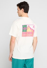 T-shirt bianco in cotone con una stampa grafica colorata sulla schiena con motivi di montagna e stelle, e il testo "ADIDAS ORIGINS" in rosa.
