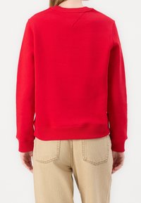 Persona vestida con un sudadera roja de manga larga lisa y pantalones beige claros, vista desde la espalda sobre un fondo blanco.