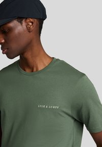 Mand iført en mørk kasket og grøn Lyle & Scott t-shirt, kigger til venstre mod en ensfarvet lys baggrund.