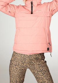 Personne portant une veste matelassée rose clair avec fermeture éclair et un pantalon imprimé léopard sur un fond uni.