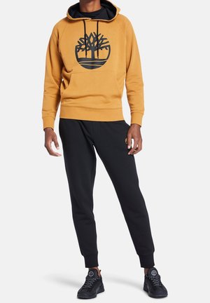 Personne portant un sweat à capuche moutarde avec un logo d'arbre noir, un pantalon de jogging noir et des baskets noires, debout sur un fond blanc uni.