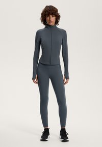 Skiefer färgad träningsjacka med hög krage och full dragkedja, parat med matchande leggings. Båda har en elegant, slät textur. Svarta träningstövlar.