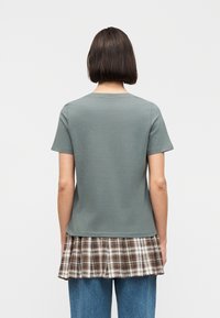 Γυναίκα με κοντά σκούρα μαλλιά που φοράει πράσινη κοντομάνικη μπλούζα πάνω από φούστα καρό σε καφέ χρώμα και μπλε τζιν, με την πλάτη στην κάμερα.