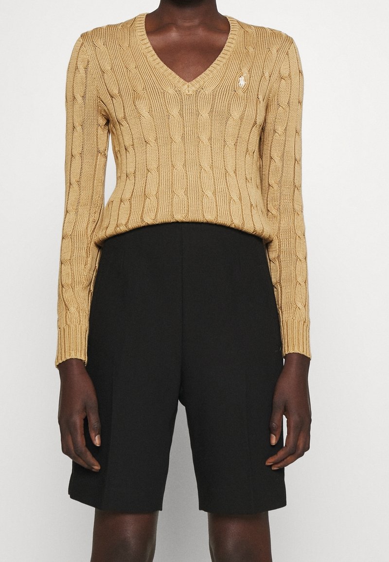 Personne portant un pull beige à col en V torsadé et un short noir au genou, debout devant un fond clair uni.