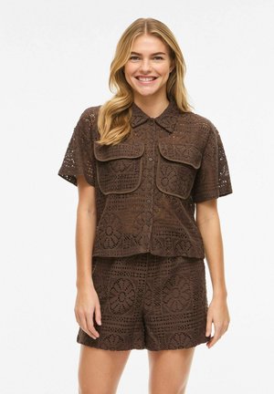 Mujer sonriente con camisa marrón de crochet de manga corta con botones y grandes bolsillos con solapa, y pantalones cortos de crochet a juego sobre fondo blanco.