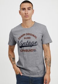 Šedé bavlněné tričko s grafickým potiskem v tmavě modré a hnědé barvě, s nápisem "DENIM ORIGINAL BLEND Vintage CPHBLND EST. 1993." Krátké rukávy.