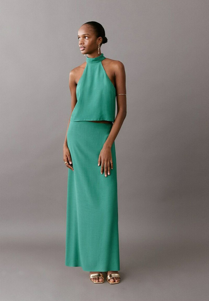 Ženska v brezrokavnem zelenem halter topu in dolgi, ujemajoči se krilu stoji ob sivem ozadju in gleda v stran.