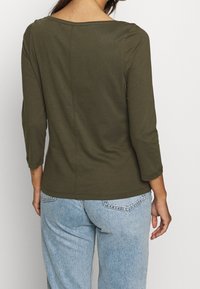 Camicia aderente a maniche lunghe verde oliva con scollatura ampia e orlo arrotondato. Il tessuto è liscio; presenta una cucitura centrale sul retro.