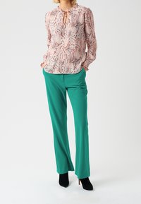 Blouse florale en rose et bleu avec un col à ouverture, associée à un pantalon vert ajusté et des bottines noires à talons. Tissu lisse, coupe décontractée.