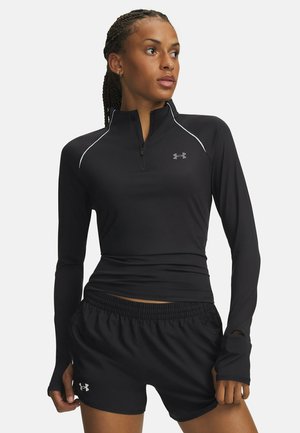 Atletische vrouw met vlechten draagt een zwart langarmig topje met rits en zwarte shorts, staat met handen nabij de heupen, kijkt naar rechts.
