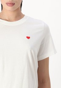 Witte katoenen t-shirt met korte mouwen, voorzien van een kleine rode hartborduursel op de linkerkant van de borst. Zachte textuur, casual ontwerp.
