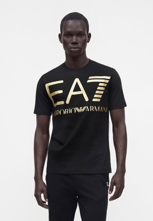 Homme portant un t-shirt noir avec un grand logo doré EA7 Emporio Armani sur le devant, assorti à un pantalon noir sur fond blanc uni.