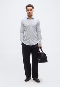 Άνδρας που φορά ανοιχτό γκρι πουκάμισο με κουμπιά και μαύρο παντελόνι, κρατώντας μια μαύρη υφασμάτινη τσάντα στο δεξί του χέρι, στέκεται μπροστά σε λευκό φόντο.