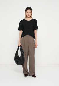 Melnas teksturētas blūzes ar puff piedurknēm, saskaņotas ar augstām brūnām biksēm. Modelis tur vaļēju melnu rokassomu un bordo apavus.