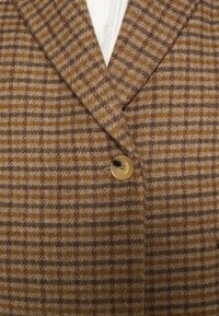 Blazer en laine à carreaux marron et beige avec un bouton unique en écaille et un col de chemise blanc visible en dessous.