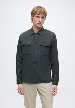 Junger Mann mit kurzen blonden Haaren, der eine dunkelgraue Hemdjacke mit zwei Brusttaschen und hellbeige Hose trägt.
