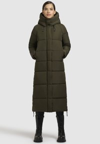 Dolga, olivno zelena puhovka z visokim kapuco, ki ima vodoravno šivane dele, stranske žepe in črne dodatke. Črne vojaške škornje.