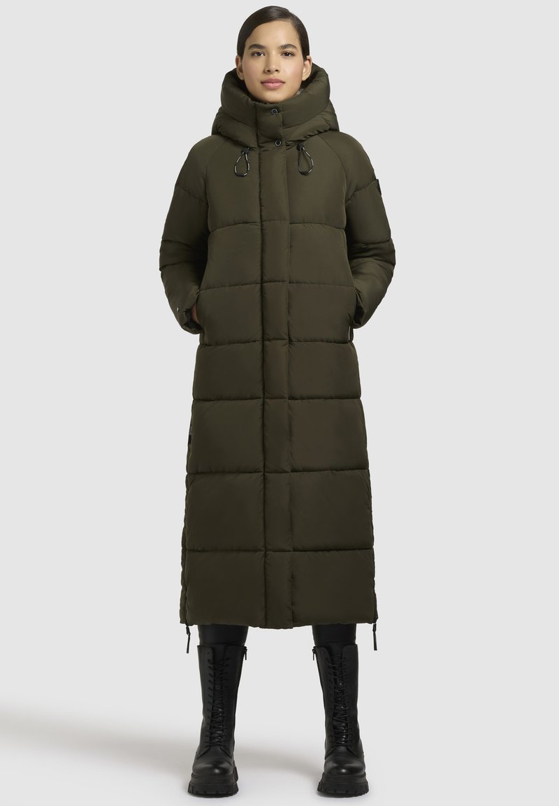 Langer, olivgrüner Daunenmantel mit hoher Kapuze, horizontal gesteppten Bereichen, Seiten Taschen und schwarzen Akzenten. Schwarze Combat-Stiefel.
