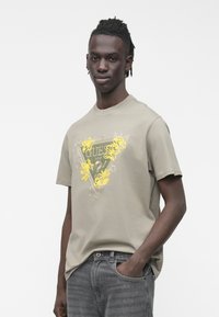 Pelēka kokvilnas T-krekls ar dzeltenu ziedu rakstu un trīsstūrveida dizainu priekšpusē. Īsas piedurknes un apaļa izgriezuma kakls.