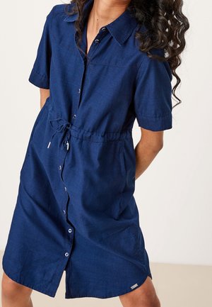 Žena stojící před neutrálním pozadím, oblečená v tmavě modrých šatech po kolena s krátkými rukávy, zapínacích na knoflíky, s límečkem a stahovacím páskem v pase.