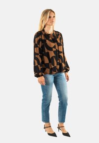 Blusa de patrones marrón y negro con mangas largas, combinada con jeans ajustados de color azul claro y zapatos de tacón alto negros.