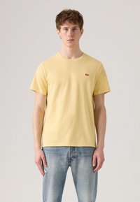 Camiseta de algodón amarilla de mangas cortas y cuello redondo, con un pequeño logotipo rojo de Levi's en el pecho. Se lleva con unos jeans desgastados de color azul claro.