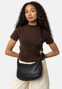 Borsa a spalla nera in pelle con forma curva e hardware dorato, impreziosita da una tracolla removibile. Modella che indossa un top in maglia marrone e pantaloni beige.