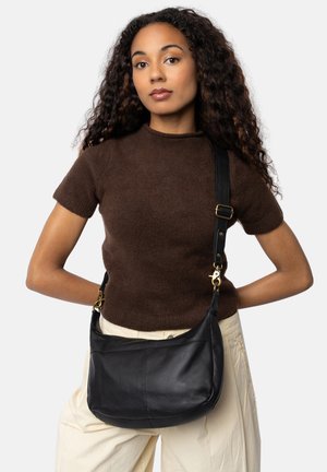 Zwarte leren schoudertas met een gebogen vorm en gouden hardware, verfraaid met een afneembare schouderband. Model draagt een bruine gebreide top en een beige broek.