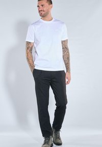 Mann mit tätowierten Armen, der ein weißes T-Shirt, schwarze Hosen und graue Schnürstiefel trägt, steht vor einem hellgrauen Hintergrund.