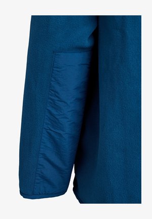 Szczegół niebieskiej kurtki polarowej, ukazujący teksturowany materiał z gładką, tkanej kieszonką. Charakteryzuje się żywym, jednolitym kolorem oraz miękką, ciepłą teksturą.