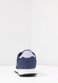 Blå sneaker med mesh och mocka material, med en vit gummisula och en lavendelaccent på hälen. "New Balance" logotyp på baksidan.