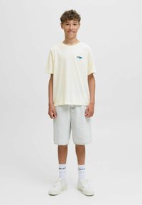 Lichtgele katoenen t-shirt met een blauw grafisch ontwerp, gecombineerd met lichtdenim shorts. Het model draagt witte sneakers en crew-sokken.