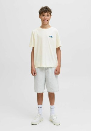 Helles, gelbes Baumwoll-T-Shirt mit einem blauen Grafikdesign, kombiniert mit hellen Jeansshorts. Das Modell trägt weiße Turnschuhe und Knöchelsocken.