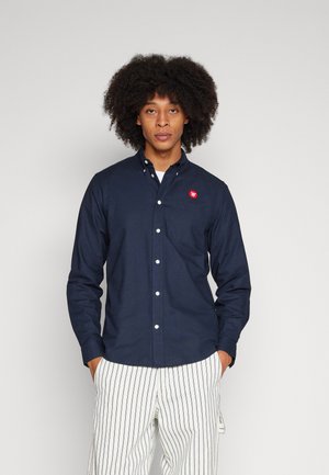 Navy button-up shirt with a chest pocket and red logo patch, paired with vertically striped light pants. Fabric appears smooth and textured.