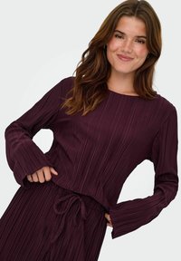 Paarse plooien jurk met lange mouwen en een ronde hals. Kenmerkt zich door een taille met trekkoord en een textuurstof voor extra detail.