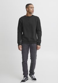 Sudadera negra con cuello redondo y un logo sutil, combinada con jeans gris oscuro y zapatillas negras con acentos en blanco.