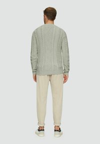 Mann, der von hinten steht, trägt einen hellgrünen, strukturierten Pullover, beigefarbene hochgekrempelte Hosen und weiße Sneakers mit schwarzen Akzenten.