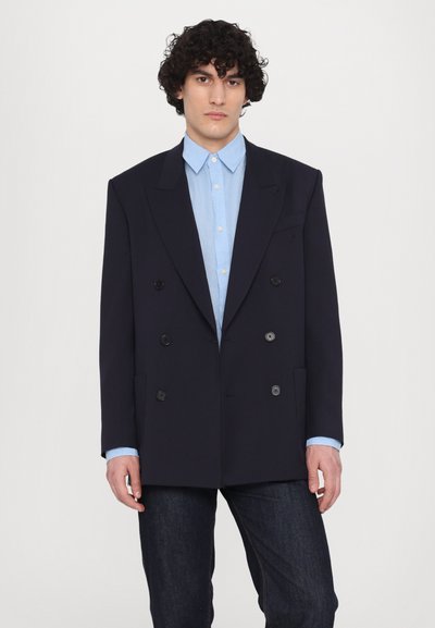Blazer bleu marine croisé à revers crantés, boutons noirs, poches avant, porté par-dessus une chemise boutonnée bleu clair et un jean noir.