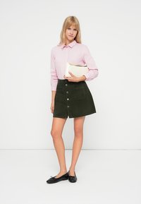 Žena v růžovém pruhovaném tričku a tmavě zelené sukni na zapínání drží krémovou kabelku, má na nohou černé baleríny, na bílém pozadí.