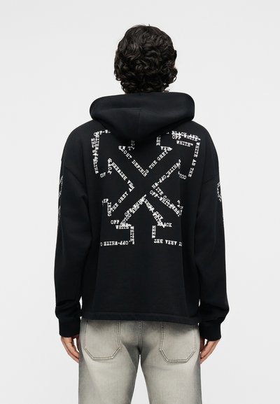 Personne aux cheveux bouclés portant un sweat à capuche noir avec un texte blanc disposé en forme de flèche dans le dos, associée à un jean gris clair.