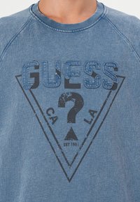 Blå sweatshirt med rund hals, med sydd "GUESS" tekst og et sort trekantet grafikk med et spørsmålstegn og årstall 1981.